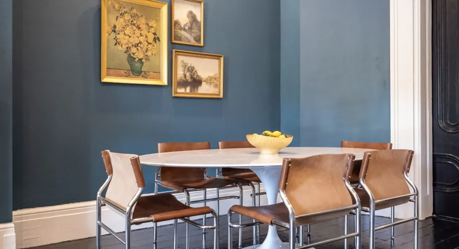 Dining table and tan chairs against a blue wall with framed artwork