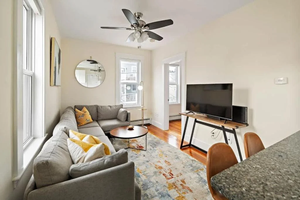 Bright living room with gray sectional sofa, yellow pillows, hardwood floors, and natural light, a quiet date spot in our